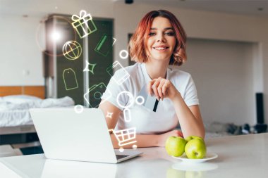 Bilgisayarlı, kredi kartlı, mutfaktaki elmalı alışveriş tabelalarıyla internetten alışveriş yapan gülümseyen bir kız. 