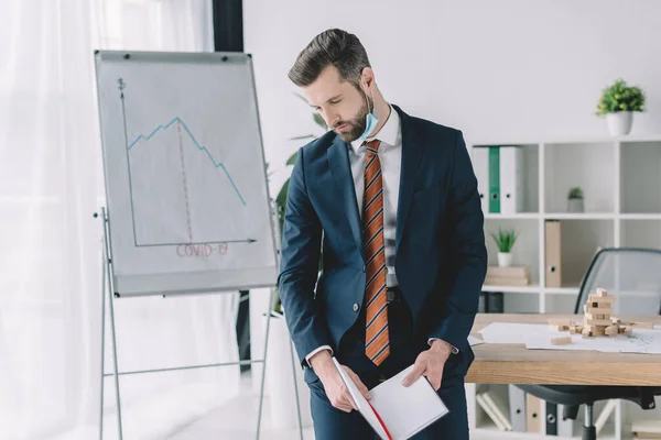 Hombre de negocios deprimido sosteniendo el cuaderno mientras está de pie con la cabeza inclinada cerca del rotafolio con gráficos que muestran disminución - foto de stock