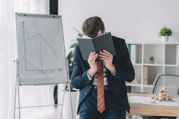 Homem de negócios deprimido cobrindo rosto com notebook enquanto estava perto flipchart com inscrição covid-19 e infográficos mostrando recessão — Fotografia de Stock