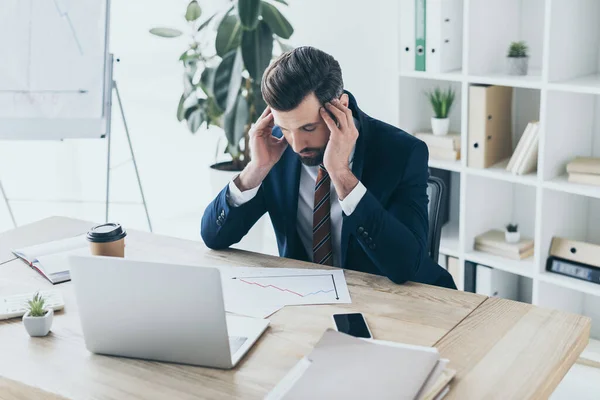 Hombre de negocios deprimido tocando la cabeza inclinada mientras estaba sentado en el lugar de trabajo con los ojos cerrados - foto de stock