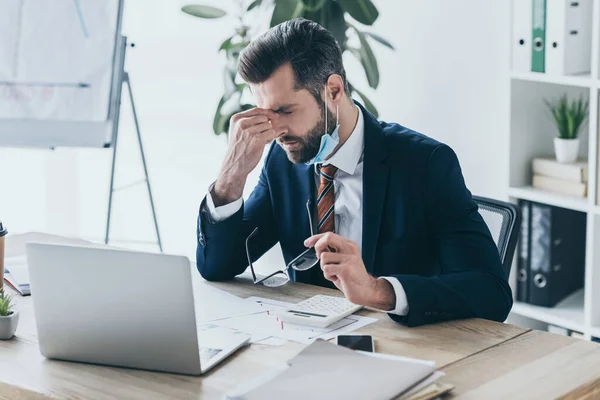 Deprimido, hombre de negocios agotado sosteniendo anteojos y tocando la cara mientras está sentado en el lugar de trabajo cerca de la computadora portátil y documentos - foto de stock