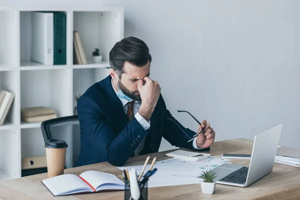 Stressed, exhausted businessman holding eyeglasses and touching face while sitting at workplace near laptop and documents — Stock Photo