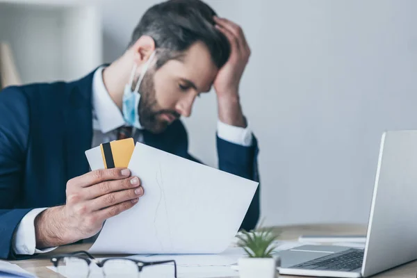 Selective focus of depressed businessman holding paper and credit cards while sitting at workplace with bowed head and closed eyes — Stock Photo