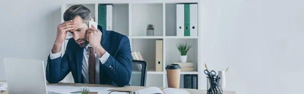 Panoramic shot of upset businessman touching forehead while talking on smartphone at workplace — Stock Photo