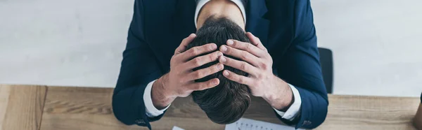 Panoramic shot of depressed businessman holding hands on bowed head while sitting at desk in office — Stock Photo