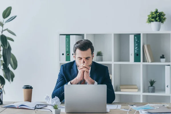 Hombre de negocios deprimido agarrado de la mano cerca de la cara mientras está sentado en el lugar de trabajo con los ojos cerrados - foto de stock