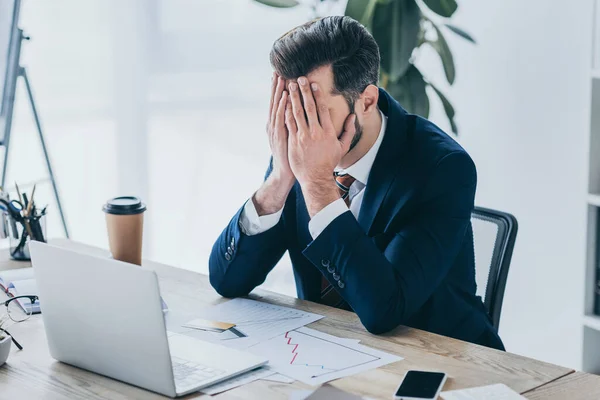 Uomo d'affari depresso che copre il viso con le mani mentre seduto sul posto di lavoro vicino al computer portatile — Foto stock