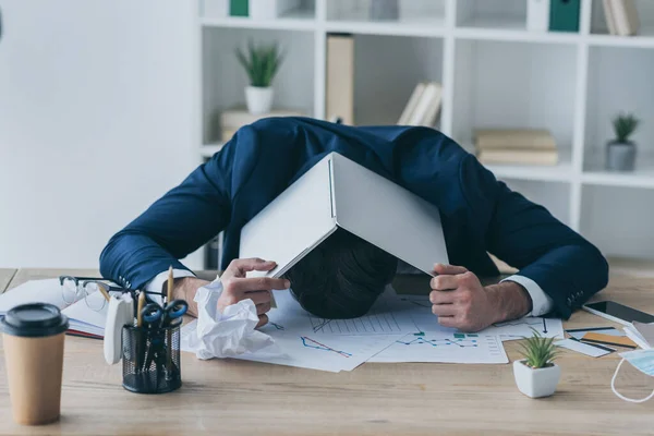 Deprimido hombre de negocios que cubre la cabeza con el ordenador portátil mientras está sentado en el lugar de trabajo - foto de stock