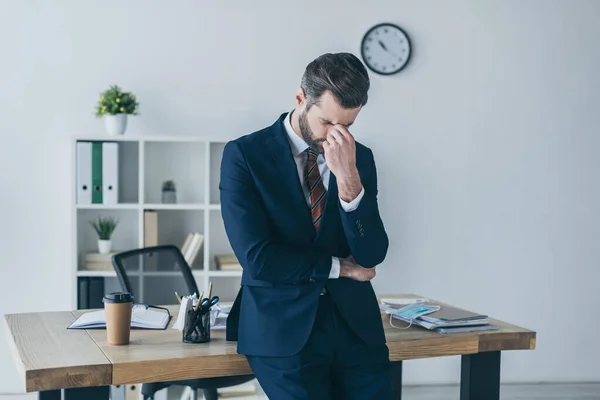 Hombre de negocios deprimido con los ojos inclinados y cerrados tocando la cara mientras está de pie en el lugar de trabajo - foto de stock