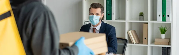 Vista cortada de homem de entrega de alimentos segurando caixas de pizza perto de empresário em máscara médica sentado no local de trabalho no escritório, tiro panorâmico — Fotografia de Stock