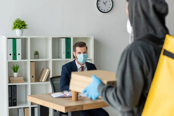 Selektiver Fokus des Essenslieferanten, der Pizzakartons in der Nähe eines Geschäftsmannes hält, der mit medizinischer Maske am Arbeitsplatz im Büro sitzt — Stockfoto