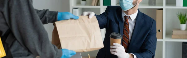 Partial view of food delivery man giving paper bag to businessman in medical mask in office, panoramic shot — Stock Photo