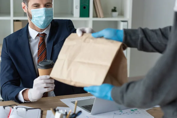 Cropped view of food delivery man giving paper bag to businessman in medical mask in office — Stock Photo