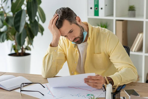 Thoughtful businessman holding hand on head while sitting at workplace and looking at document — Stock Photo