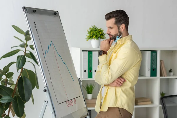 Nachdenklicher Geschäftsmann berührt Kinn beim Betrachten von Flipchart mit Covid-19-Beschriftung und Grafiken, die Rezession zeigen — Stockfoto