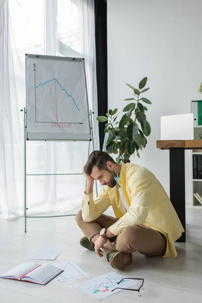 Hombre de negocios deprimido tocando la cabeza mientras está sentado en el suelo cerca de documentos y rotafolio con inscripción covid-19 y gráficos que muestran la recesión - foto de stock