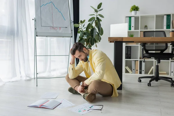 Depressed businessman sitting on floor near papers, notebook and flipchart with covid-19 inscription and graphs showing decrease — Stock Photo