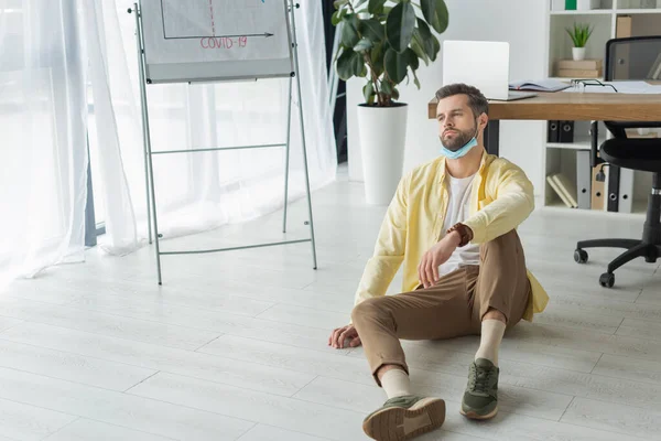 Depressed businessman sitting on floor near flipchart with covid-19 inscription and looking away — Stock Photo