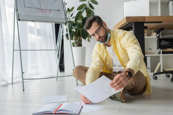 Serious, thoughtful businessman looking at paper while sitting on floor near flipchart with covid-19 inscription — Stock Photo