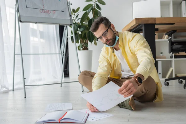 Hombre de negocios serio y reflexivo mirando el papel mientras está sentado en el piso cerca del rotafolio con la inscripción covid-19 - foto de stock