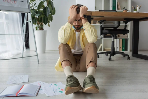 Homme d'affaires déprimé tenant la main sur la tête tout en étant assis sur le sol avec les yeux fermés près d'un ordinateur portable et des papiers avec des graphiques — Photo de stock