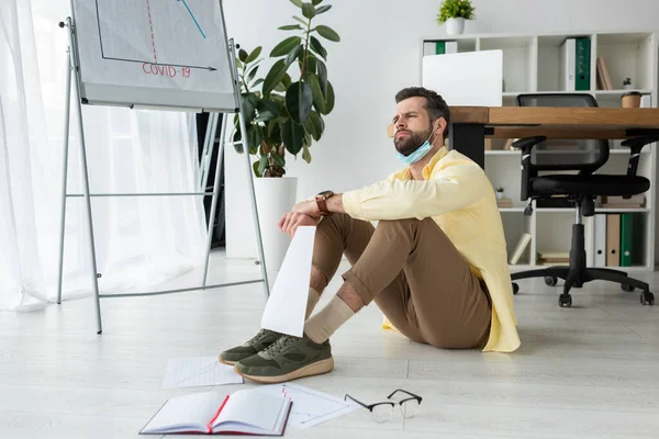 Uomo d'affari premuroso seduto sul pavimento e guardando lontano vicino a carte, quaderno e lavagna a fogli mobili con iscrizione covid-19 e infografica — Foto stock