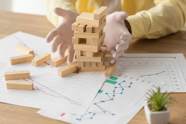 Vista recortada de hombre de negocios en guantes de látex cerca de caer bloques de madera torre y papeles con infografías - foto de stock