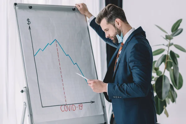 Uomo d'affari premuroso guardando carta mentre in piedi vicino flipchart con iscrizione covid-19 e grafici che mostrano diminuzione — Foto stock