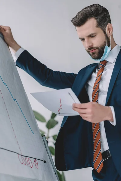 Thoughtful businessman looking at document while standing near flipchart with covid-19 inscription and graphs showing decrease — Stock Photo
