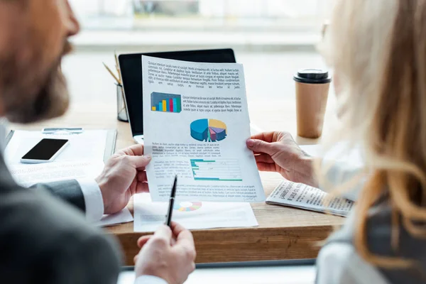 Enfoque selectivo de la gente de negocios sosteniendo papel con gráficos cerca de gadgets y taza de papel en la mesa - foto de stock