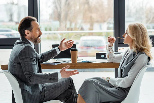 Vue latérale d'un bel homme d'affaires parlant avec une femme d'affaires près d'une tasse en papier et des documents sur la table — Photo de stock
