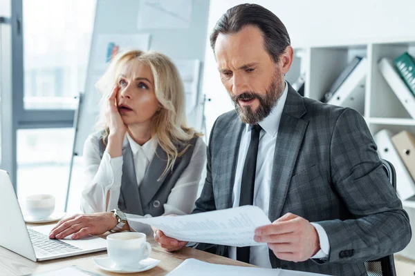 Handsome businessman holding document near thoughtful businesswoman using laptop at table in office — Stock Photo