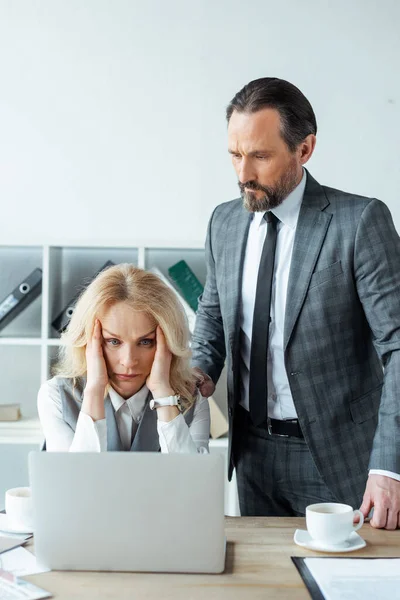 Красивый бизнесмен, стоящий рядом с концентрированной бизнесвумен глядя на ноутбук в офисе — стоковое фото