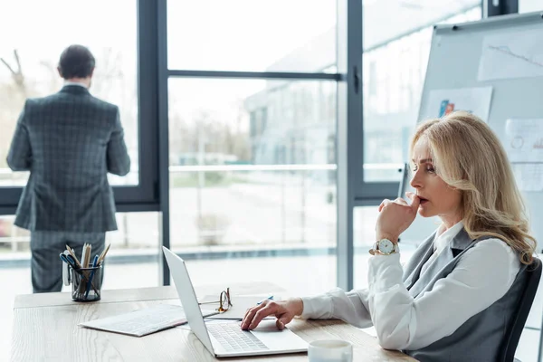 Focus selettivo della donna d'affari pensosa che utilizza il computer portatile mentre l'uomo d'affari si trova vicino alla finestra in ufficio — Foto stock