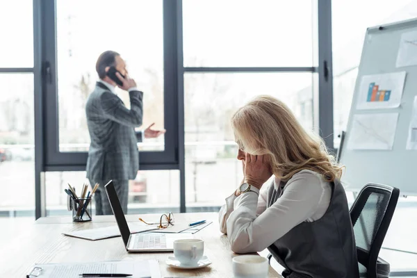 Seitenansicht einer Geschäftsfrau, die mit Laptop und Papieren am Tisch sitzt, während ein Geschäftsmann im Büro auf dem Smartphone spricht — Stockfoto