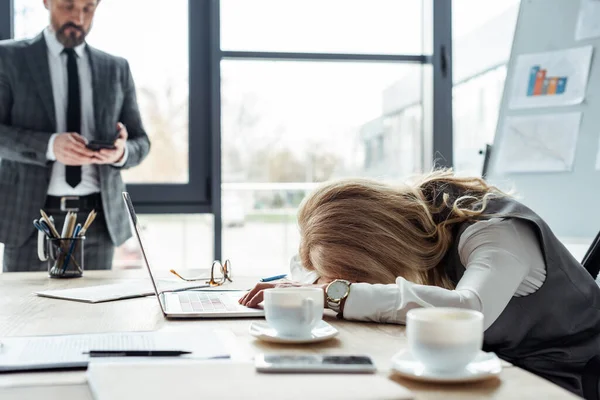 Selective focus of tired businesswoman lying on table near laptop and coffee cups while businessman using smartphone in office — Stock Photo