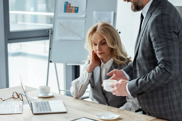 Businessman holding coffee cup near businesswoman and laptop on table in office — Stock Photo