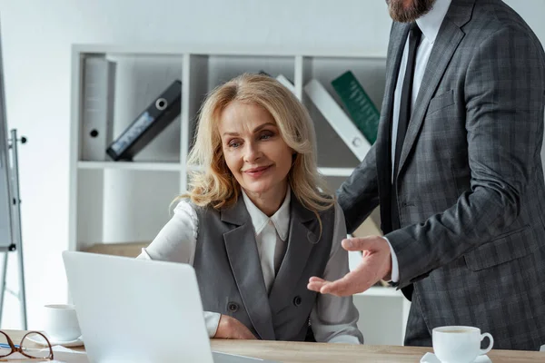 Empresário apontando com a mão perto sorridente empresária olhando para laptop na mesa no escritório — Fotografia de Stock