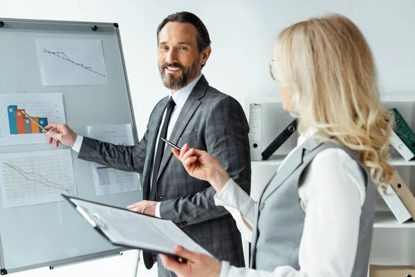 Selektiver Fokus eines lächelnden Geschäftsmannes, der auf Diagramme auf Whiteboard zeigt, in der Nähe einer Geschäftsfrau, die Klemmbrett im Büro hält — Stockfoto