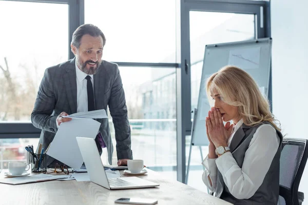 Selektiver Fokus einer Geschäftsfrau mit gebetsmühlenartig aussehendem Laptop in der Nähe eines lächelnden Geschäftsmannes, der Papiere im Büro hält — Stockfoto