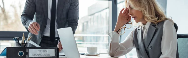 Panoramaaufnahme einer Geschäftsfrau, die auf Laptop neben Ordner mit Krisenmanagement und Geschäftsmann im Büro blickt — Stockfoto