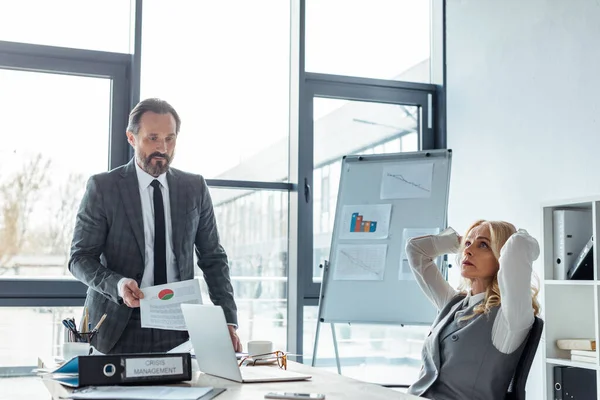 Concentration sélective d'une femme d'affaires réfléchie assise à table près d'un homme d'affaires tenant du papier avec un tableau dans son bureau — Photo de stock