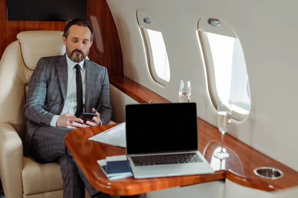 Enfoque selectivo del hombre de negocios utilizando el teléfono inteligente cerca de la computadora portátil y champán en avión - foto de stock