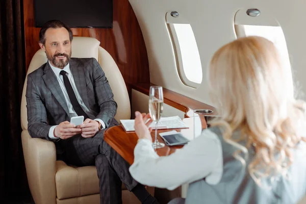 Selektiver Fokus lächelnder Geschäftsleute mit Smartphone, die Geschäftsfrau mit Champagner im Flugzeug anschauen — Stockfoto