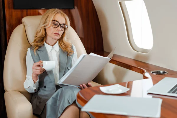 Selektiver Fokus der Geschäftsfrau mit Tasse Kaffee beim Blick auf Papiere im Flugzeug — Stockfoto