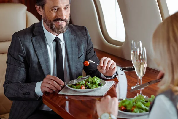 Foco seletivo de homem de negócios sorrindo comendo salada perto de empresária e champanhe no avião — Fotografia de Stock