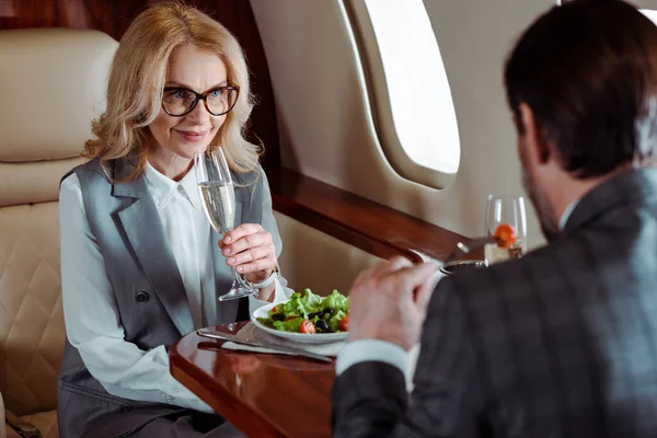 Foco seletivo de mulher de negócios sorridente segurando copo de champanhe perto de homem de negócios comendo salada no avião — Fotografia de Stock