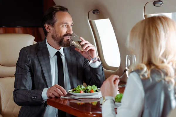 Foco seletivo de homem de negócios sorrindo bebendo champanhe perto de empresária comendo salada no avião — Fotografia de Stock