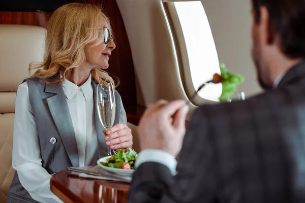 Foco seletivo de empresária segurando copo de champanhe perto de empresário comendo salada no avião — Fotografia de Stock