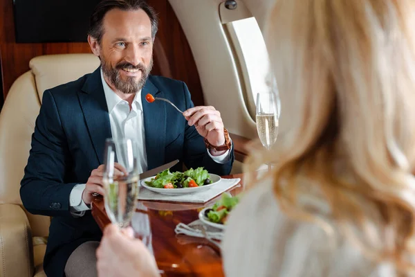 Selektiver Fokus eines lächelnden Geschäftsmannes, der Salat in der Nähe einer Geschäftsfrau mit einem Glas Champagner im Flugzeug isst — Stockfoto